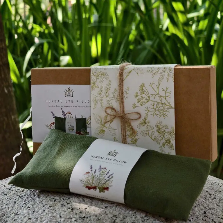 Gối chườm mắt thảo mộc màu xanh rêu cùng hộp kraft thắt dây cói, đặt trên đá trước khóm lá